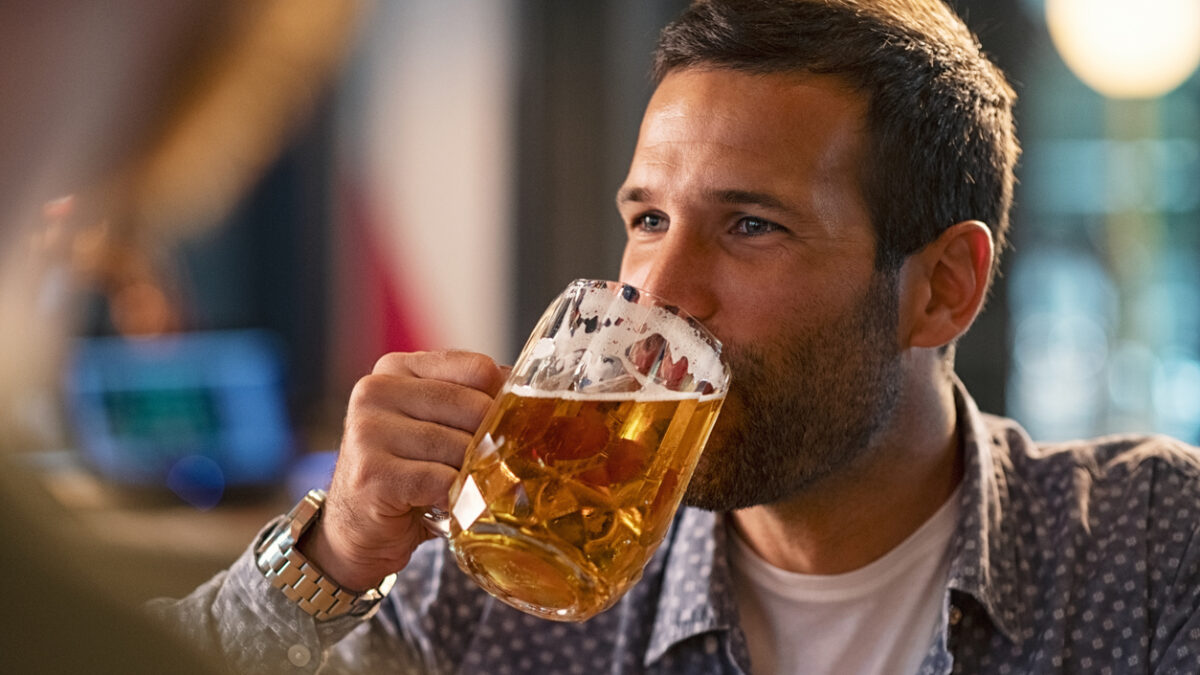 Handsome young man drinking a pint of beer while looking away. Cheerful mid man in casual clothing feeling relaxed while enjoying draft beer in bar. Middle aged guy take a sip from his drink at pub during the night.
