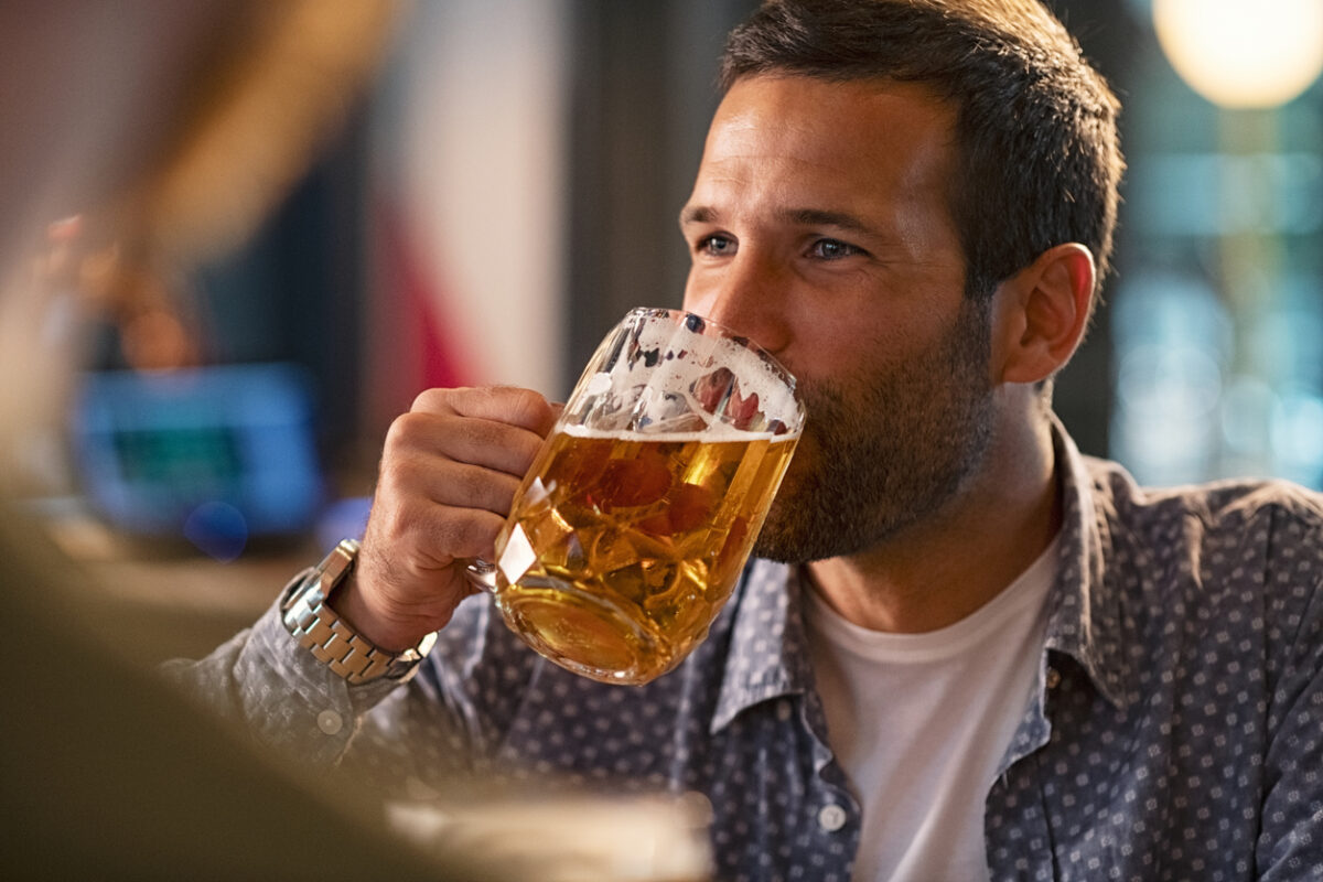 Handsome young man drinking a pint of beer while looking away. Cheerful mid man in casual clothing feeling relaxed while enjoying draft beer in bar. Middle aged guy take a sip from his drink at pub during the night.