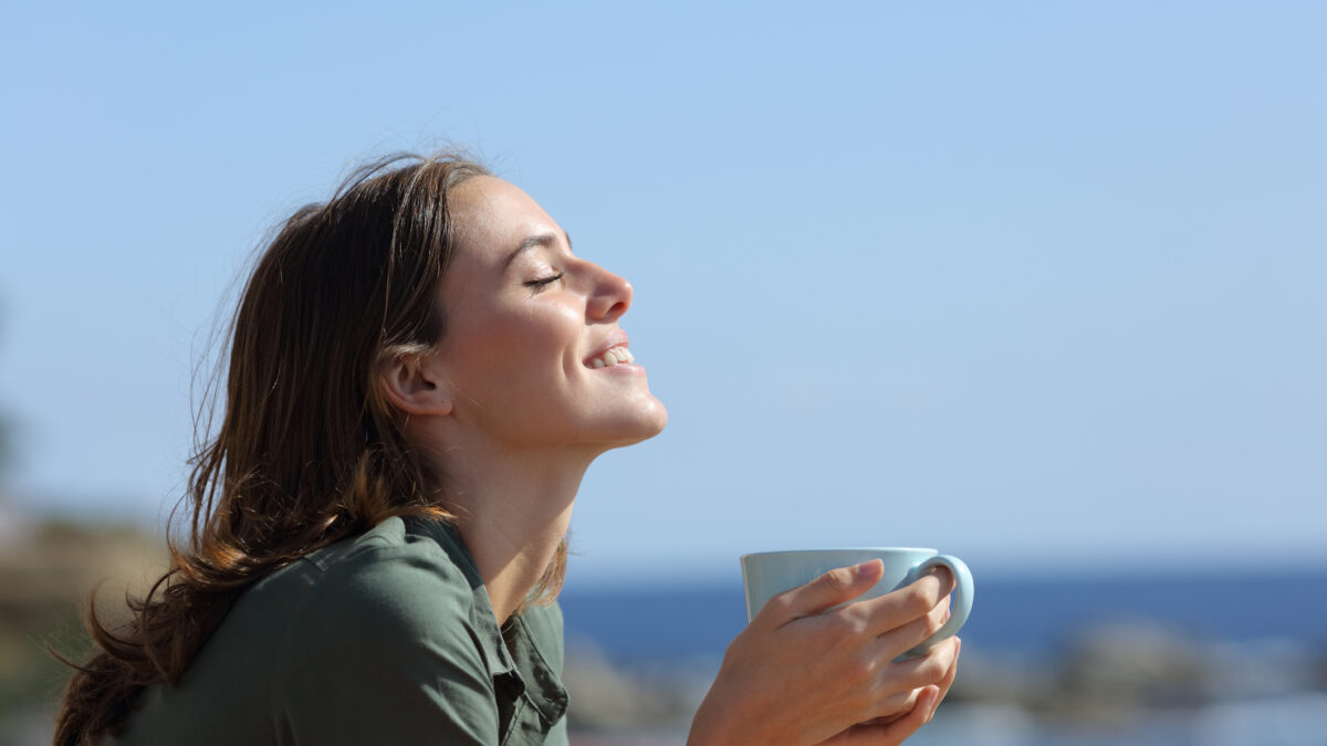 Happy woman holding coffee mug breaths on the beach