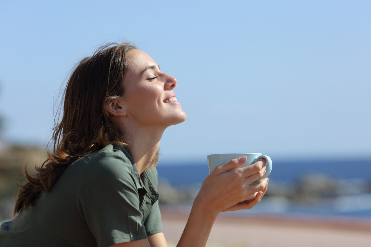 Happy woman holding coffee mug breaths on the beach