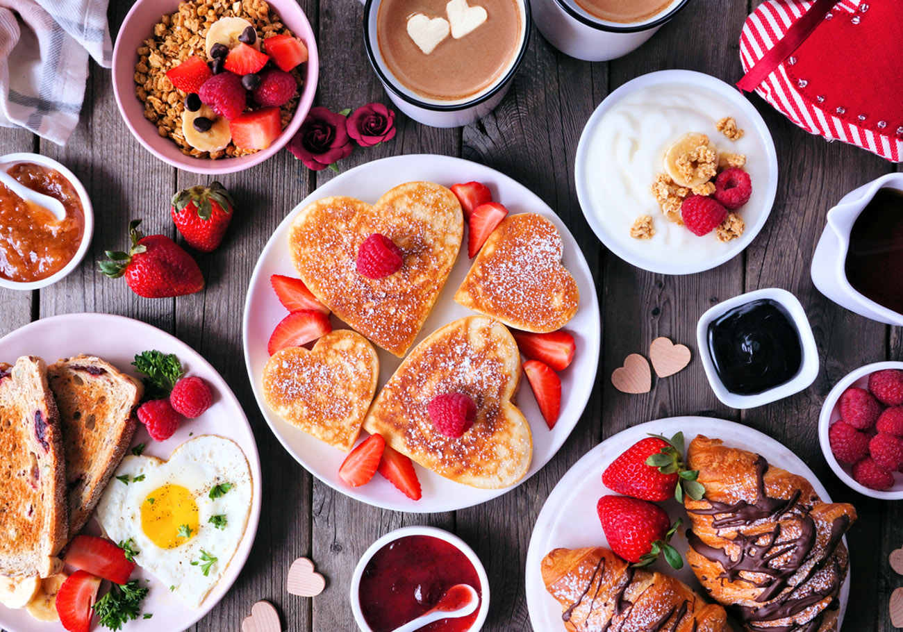 Valentines or Mothers Day brunch table scene. Top view on a dark wood background. Heart shaped pancakes, eggs and an assortment of love themed food.