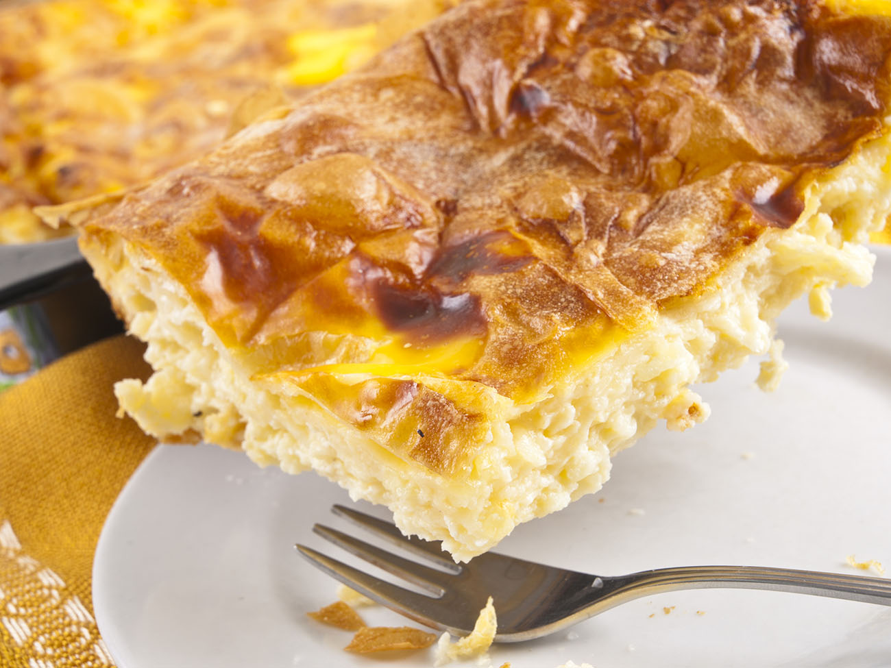 Home made cheese pie in baking tray, one piece on white plate with fork. Yellow tablecloth.