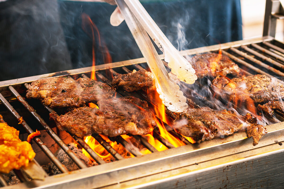 A pair of serving tongs expertly turning sizzling steaks on a lively grill. Thick tongues of flame rise dramatically from the glowing white charcoal below, illuminating the juicy meat and adding a dynamic, appetizing feel to the scene. The grill and tongs are stained with fat and oil from cooking.