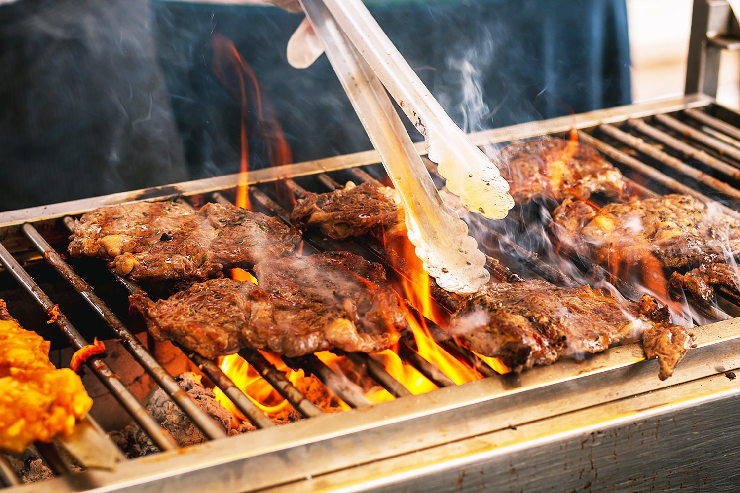 A pair of serving tongs expertly turning sizzling steaks on a lively grill. Thick tongues of flame rise dramatically from the glowing white charcoal below, illuminating the juicy meat and adding a dynamic, appetizing feel to the scene. The grill and tongs are stained with fat and oil from cooking.