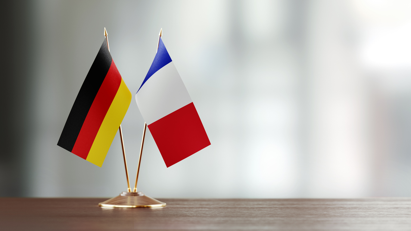 German and French flag pair on desk over defocused background. Horizontal composition with copy space and selective focus.
