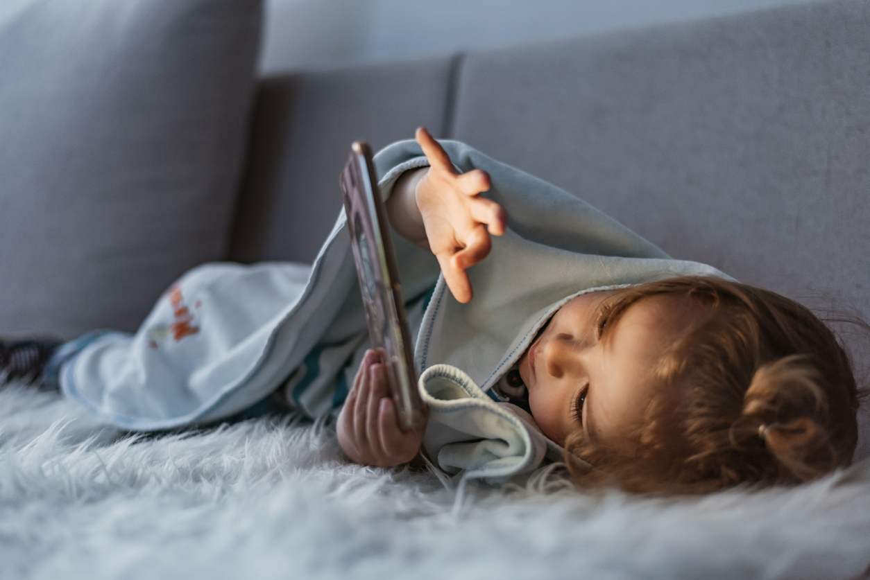 Close up of adorable kid laying down on the bed by holding a smartphone at night. Photo of Little kid in bed under a blanket looking at the smartphone at night. Happy smiling baby boy laying on his side on the bed under a blanket and plays on a smartphone in a game in the dark. The child's face is illuminated by a bright monitor.