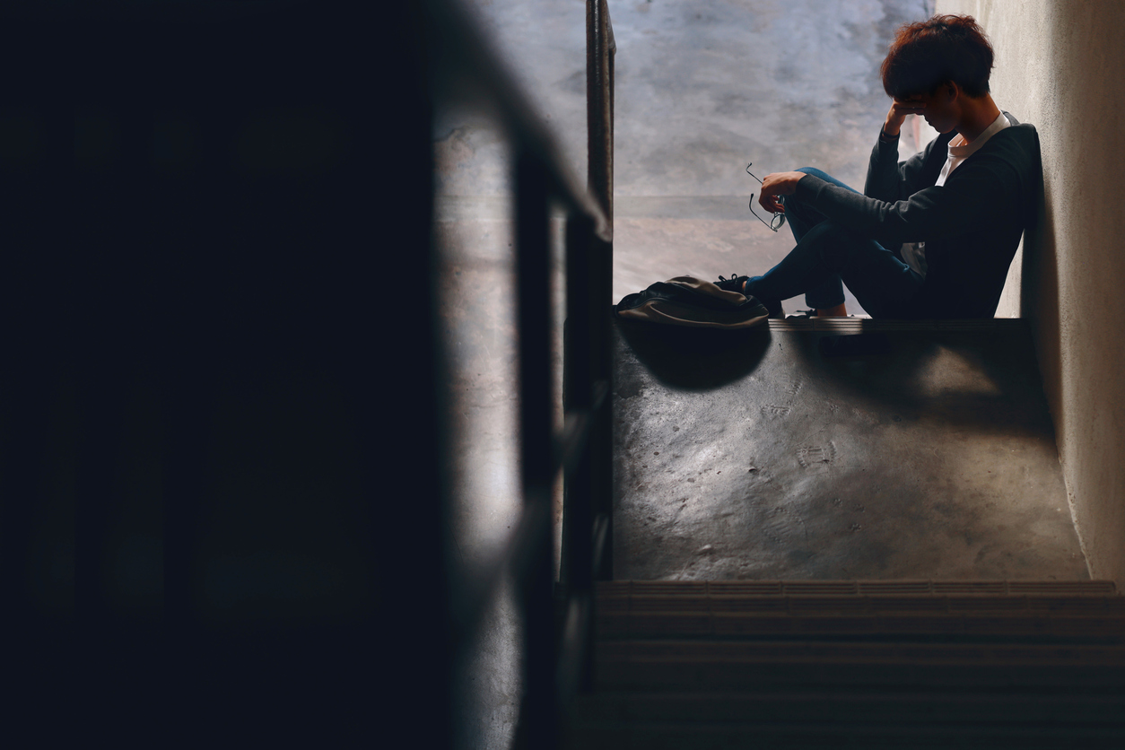 An upset Asian teenage boy student, dressed in casual outfit, sits on a stairway reading on his smartphone with a backpack beside him, while dealing with the issue of cyberbullying.