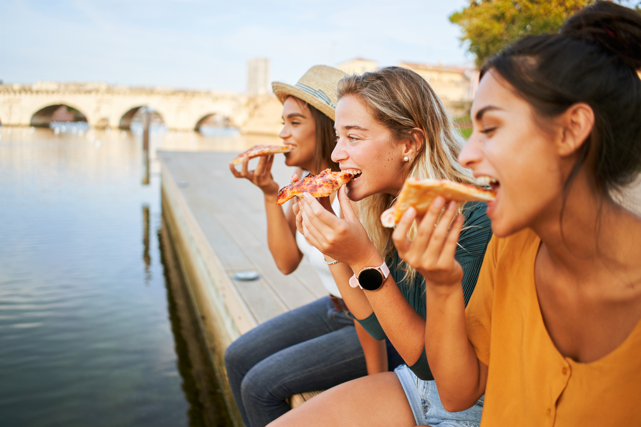 Three beautiful girls eat pizza on the lakeside. Group of women enjoy free time together sightseeing in a roman city in europe. High quality photo