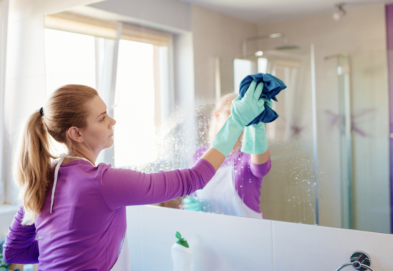 Young beautiful woman cleaning mirror in bathroom. Cleaning service. Maid cleaning at home