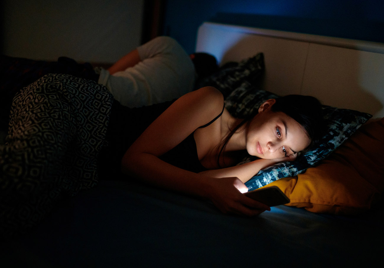 Young woman sending text message as her boyfriend sleeps in the bed, dark, copy space