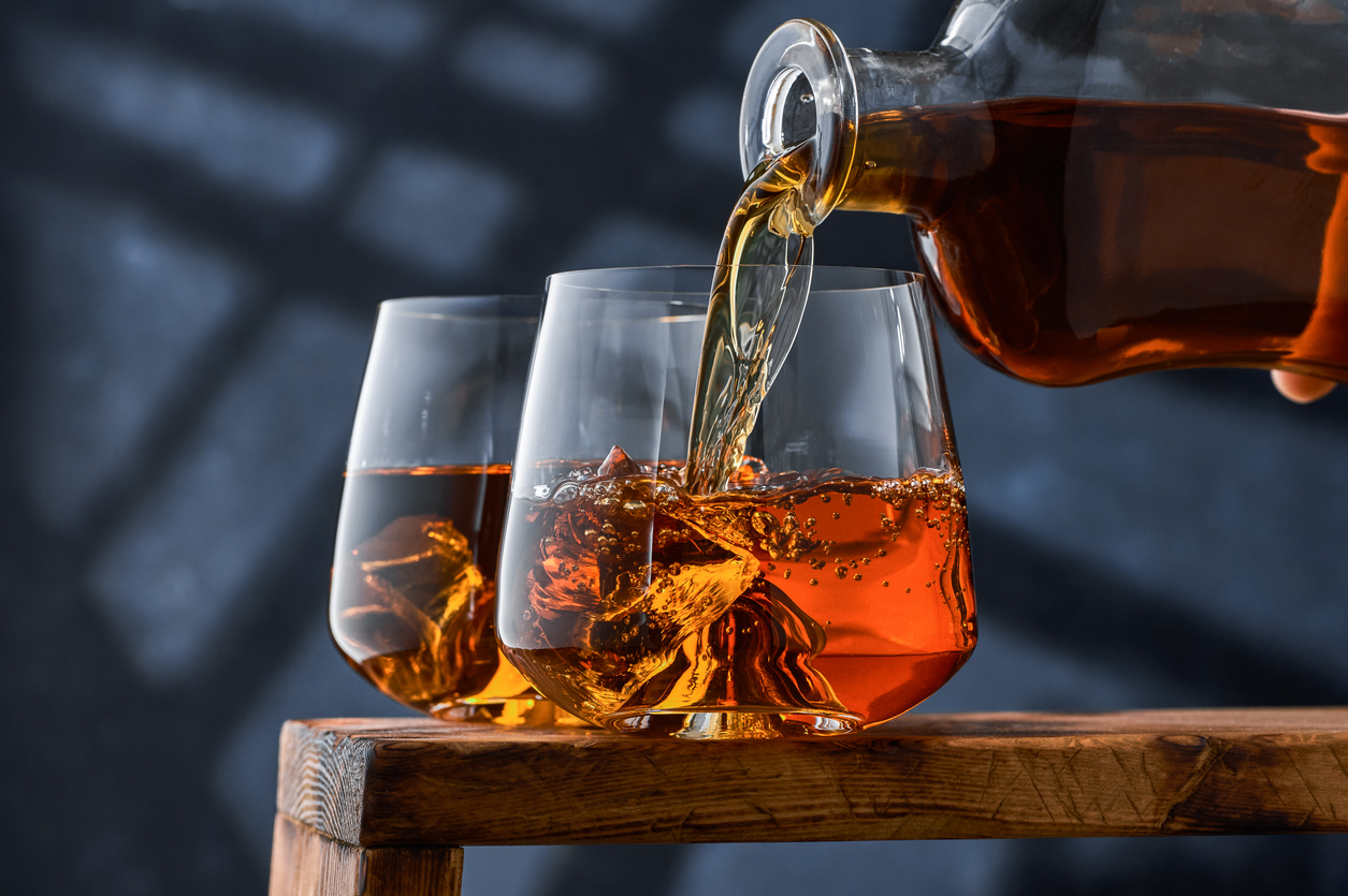 Two glasses of whiskey with ice on the counter, the glass is filled with whiskey from the bottle. Two glasses of brandy on a tray on a blue background, the glass is filled with brandy from the bottle.