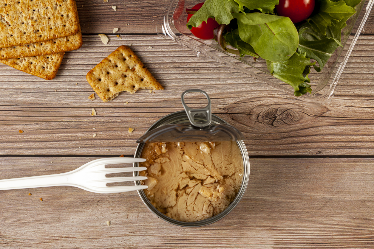 Flat lay image of quick and snack photo featuring an easy open can of tuna with tuna chunks in oil on wooden table or bench, clear plastic container with salad, crackers and a disposable plastic fork.
