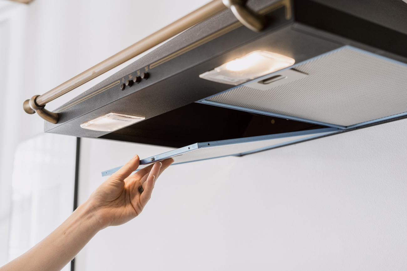 cropped shot of black metal hood extractor and person hand taking aluminum mesh filter on white wall background, maintenance household appliances