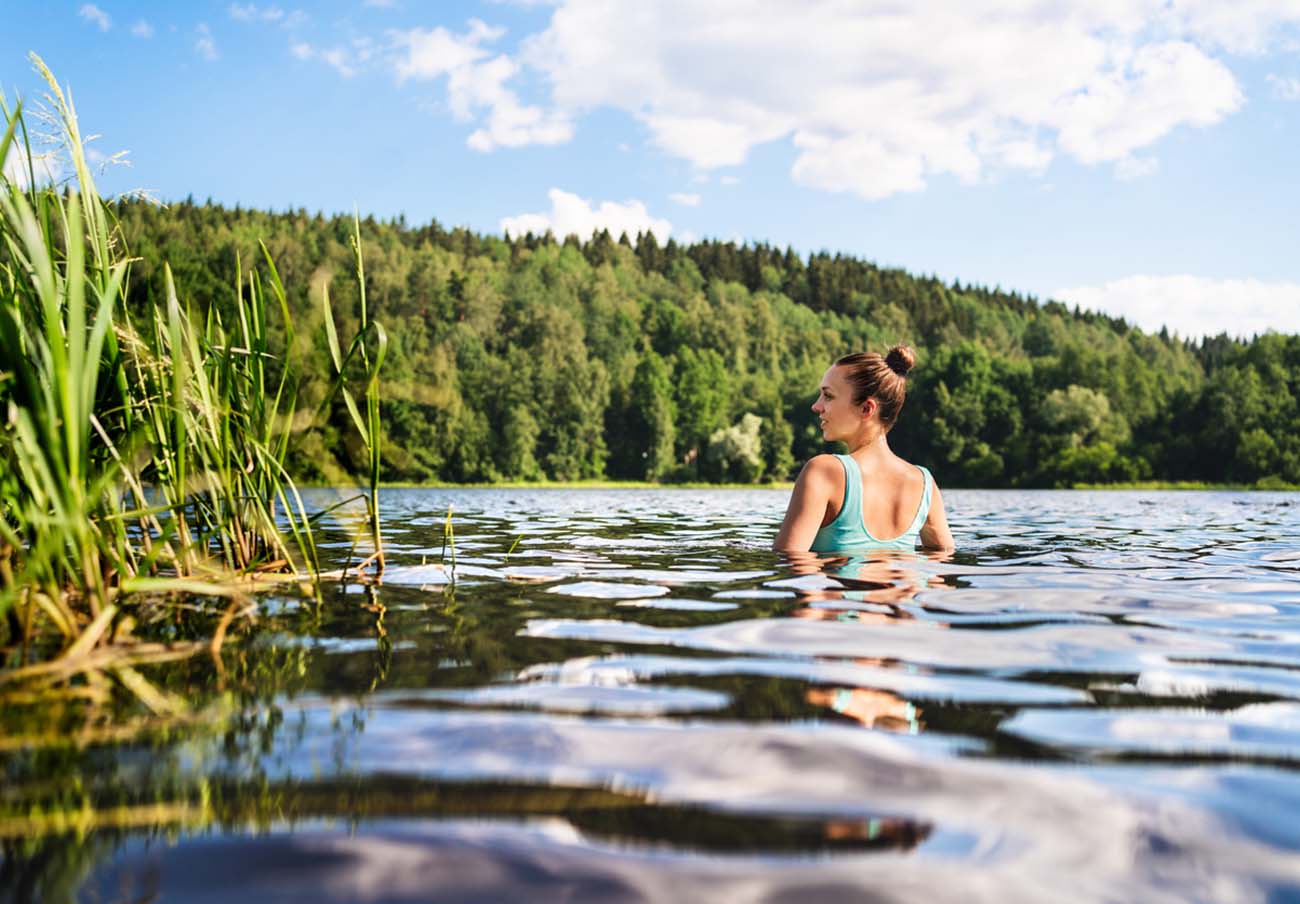 Swimming in lake in Finland. Woman in summer at beach. Finnish bathing in water in nature. Back view of person. Blue sky and beautiful green forest. Relax in Scandinavia after cottage sauna.