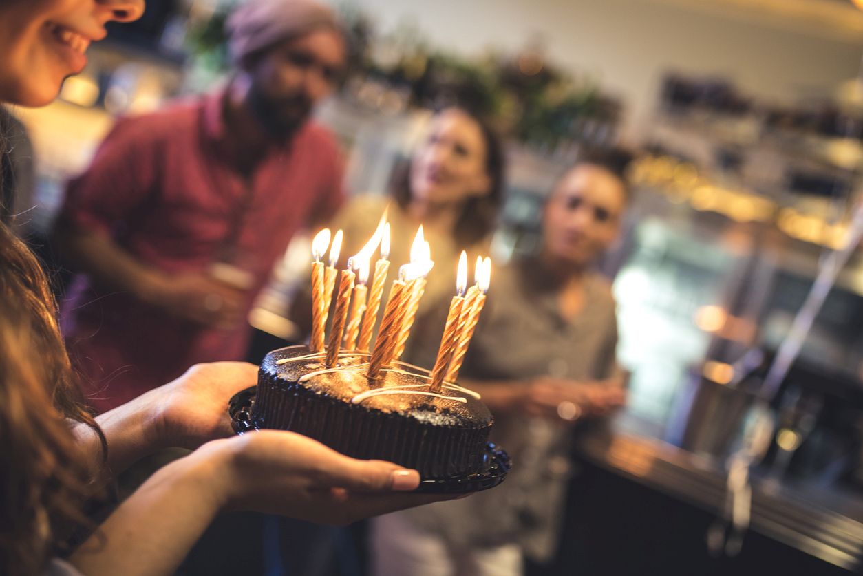 Young woman holding a birthday cake in a bar. She is ready to blow the candles. Her friends standing in back, at bar counter, and they are defocused.
