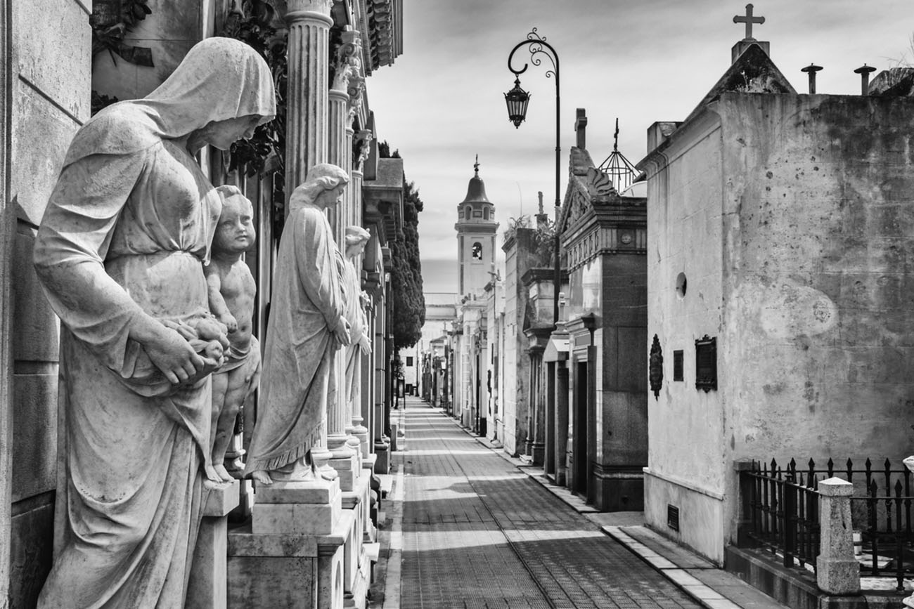 Black and white photo of an alley with mausoleums at the landmark La Recoleta Cemetery in Buenos Aires, Argentina.