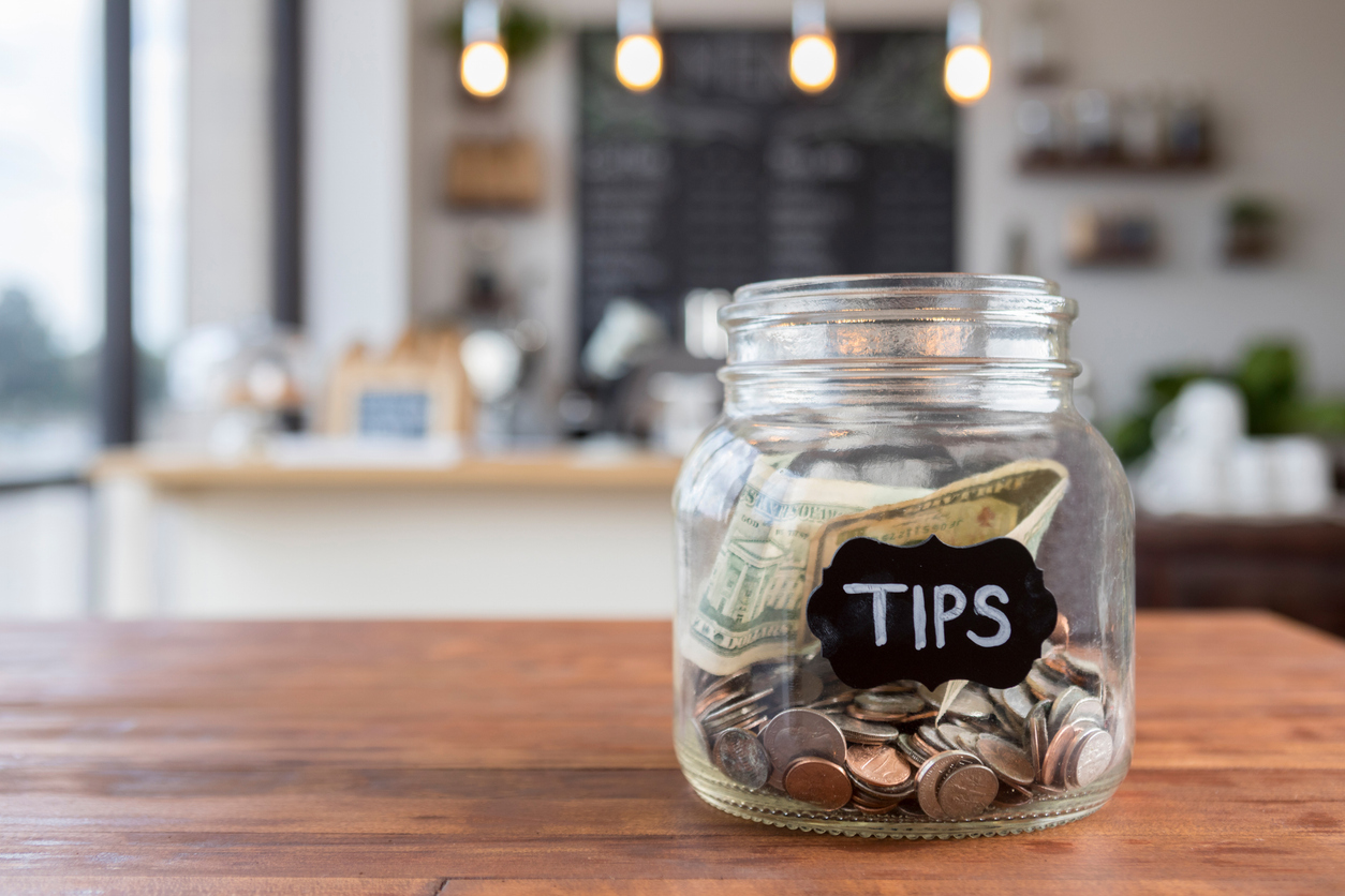 Foreground focus is on a glass jar labeled "tips" in chalk.  The jar is sitting to one side on a rustic wooden table, full of coins and bills, with coffee shop scenery in the background.