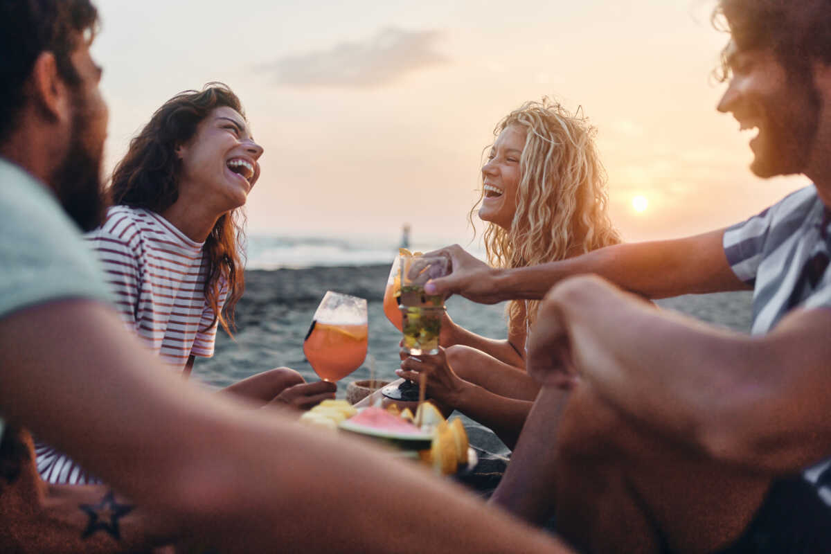 Group of cheerful friends talking while having fresh fruit and cocktails during summer sunset on the beach. Copy space.
