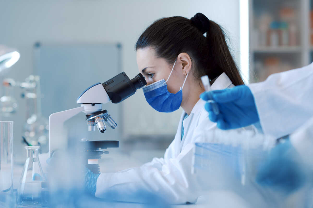 Young female researcher working in the science lab, she is wearing a face mask and using a microscope