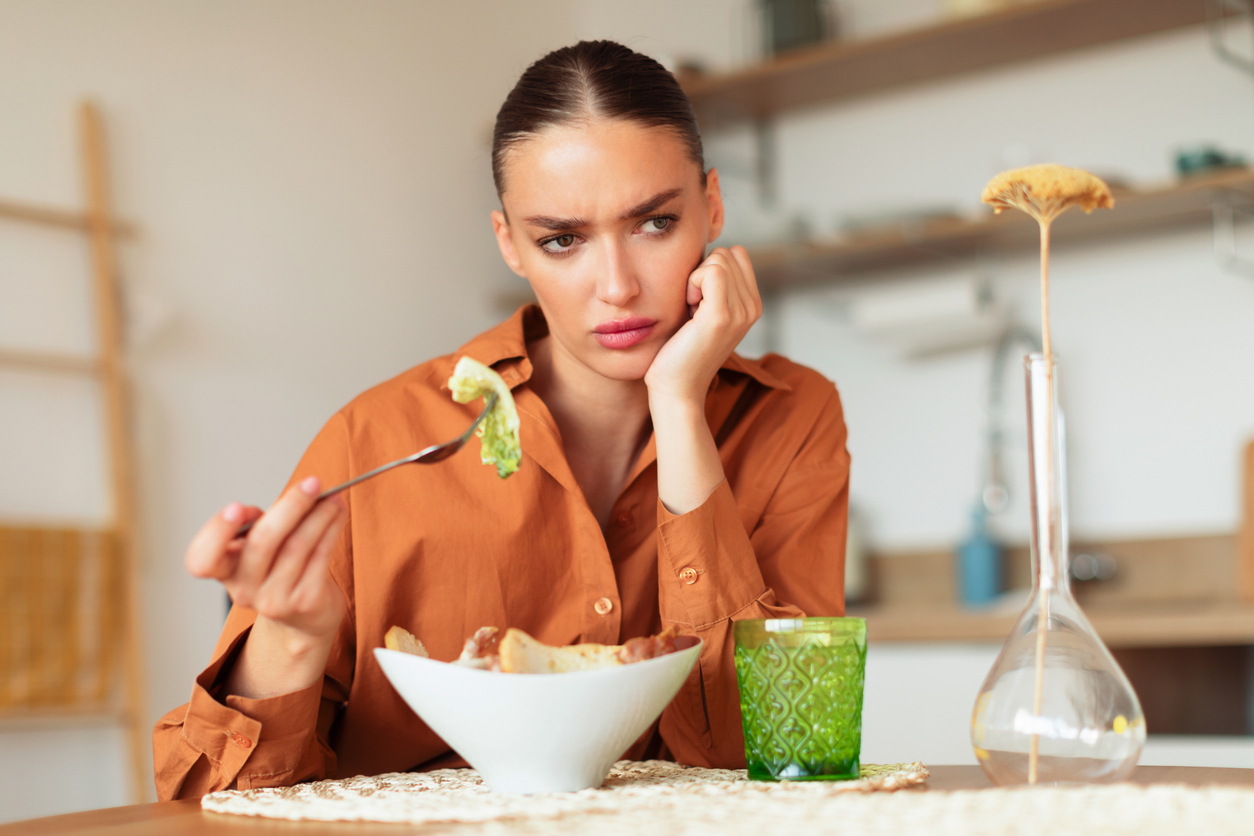 Thoughtful young woman eating caesar salad and having lack of appetite, sitting at table in kitchen interior, copy space. Digestive problems as well as spoiled and tasteless food