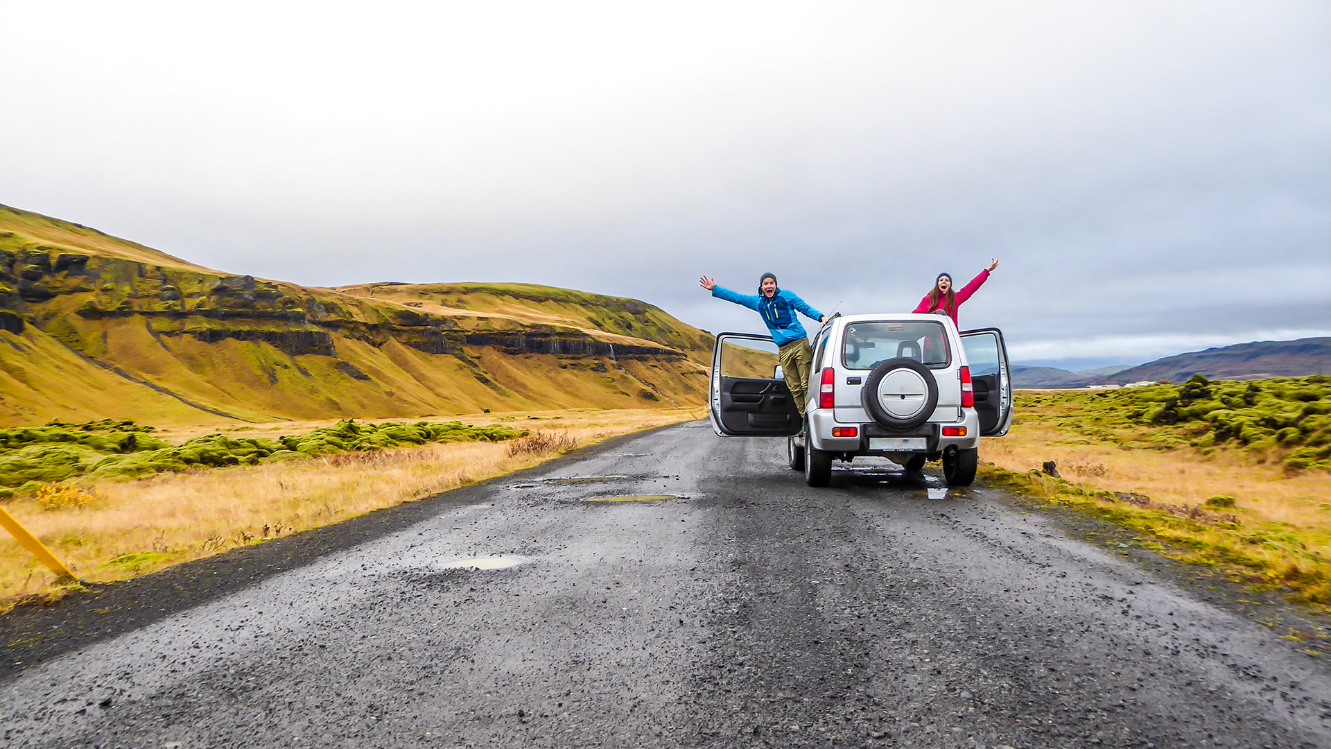 A couple stretching from the sides of the car and waving at the camera. The car is pulled on the side of the road, next to mountains. Mountains are overgrown with yellow and green grass. Fun moments