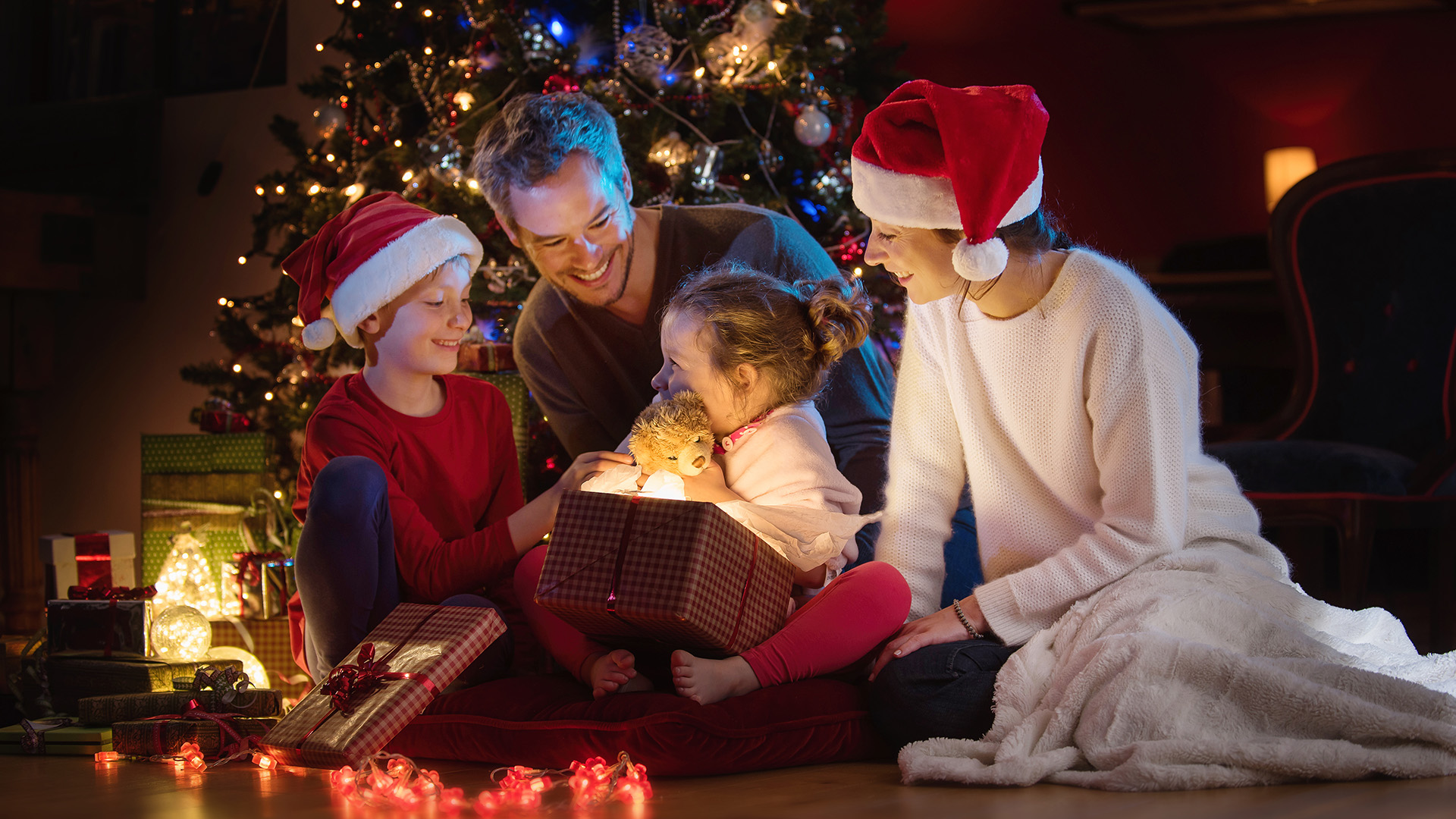 Christmas night. Near the christmas tree a lovely family opening their gifts. They enjoy the warm Christmas atmosphere in their living room, mom and kids wearing a hat of Santa Claus.