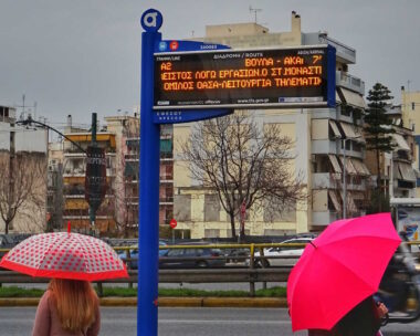 Αθηναίοι περιμένουν σε στάση λεωφορείου με την ηλεκτρονική ένδειξη να αναγράφει τον χρόνο άφιξης του μέσου μαζικής μεταφοράς