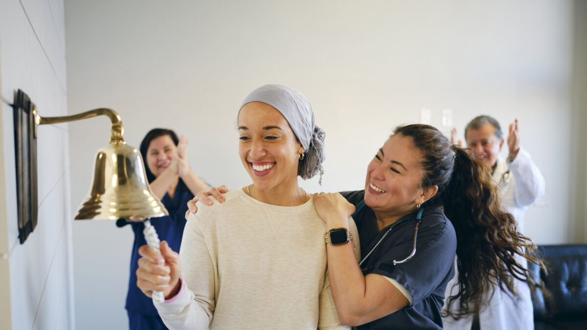 An adult woman chemotherapy patient in a treatment office, celebrating the completion of her treatment with a ceremonial bell ring. Actor portrayal