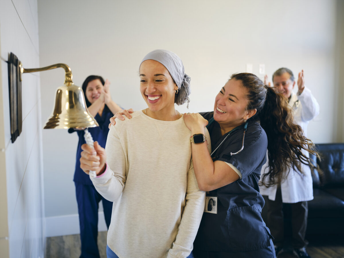 An adult woman chemotherapy patient in a treatment office, celebrating the completion of her treatment with a ceremonial bell ring. Actor portrayal
