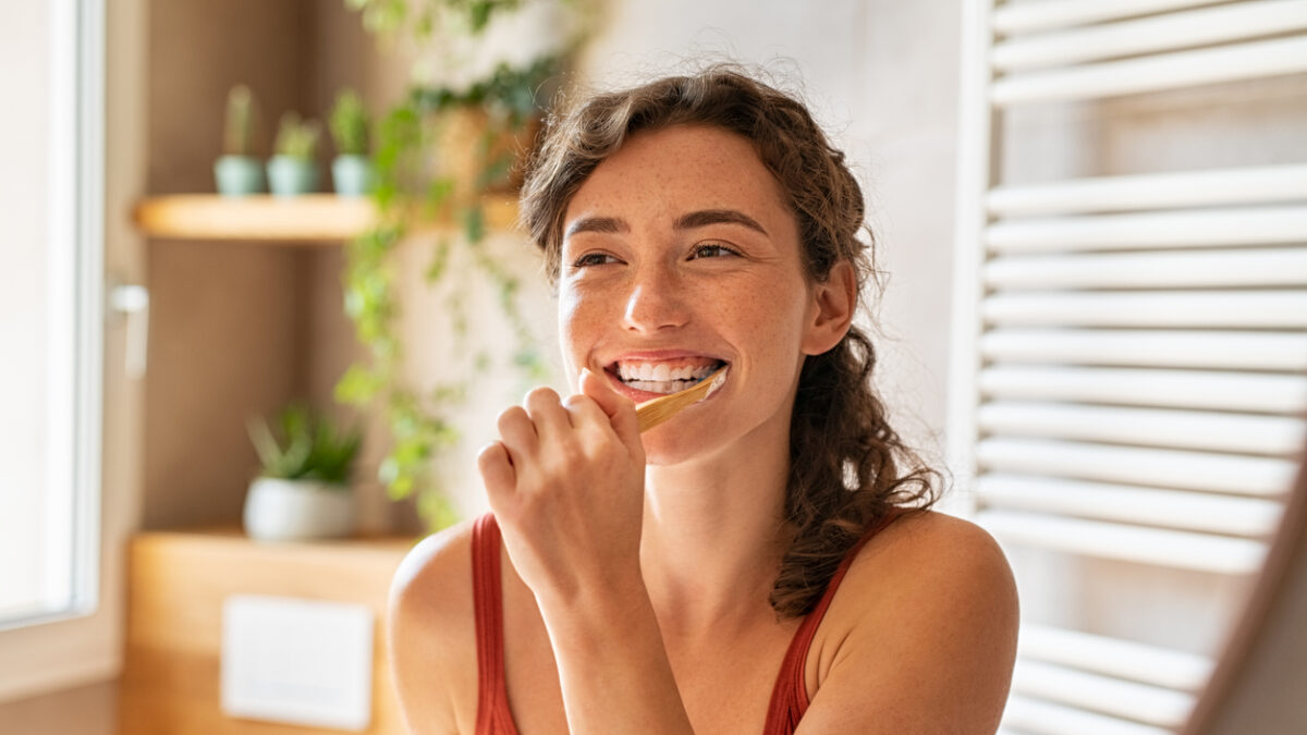 Smiling young woman brushing teeth in bathroom. Portrait of happy girl looking in mirror while using ecological toothbrush with whitening toothpaste. Beauty girl in bathroom cleaning teeth in the morning time.