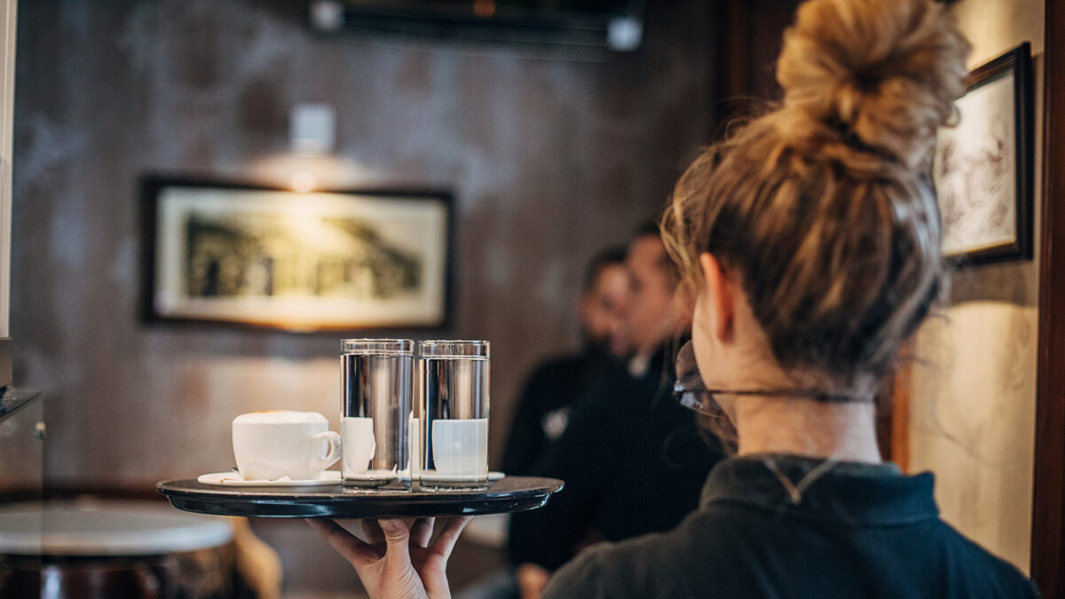 Group of people, female waitress working at cafe, wearing a protective mask. She is holding a trey with coffee cups