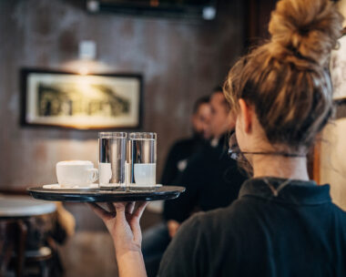 Group of people, female waitress working at cafe, wearing a protective mask. She is holding a trey with coffee cups