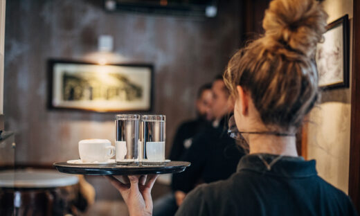 Group of people, female waitress working at cafe, wearing a protective mask. She is holding a trey with coffee cups