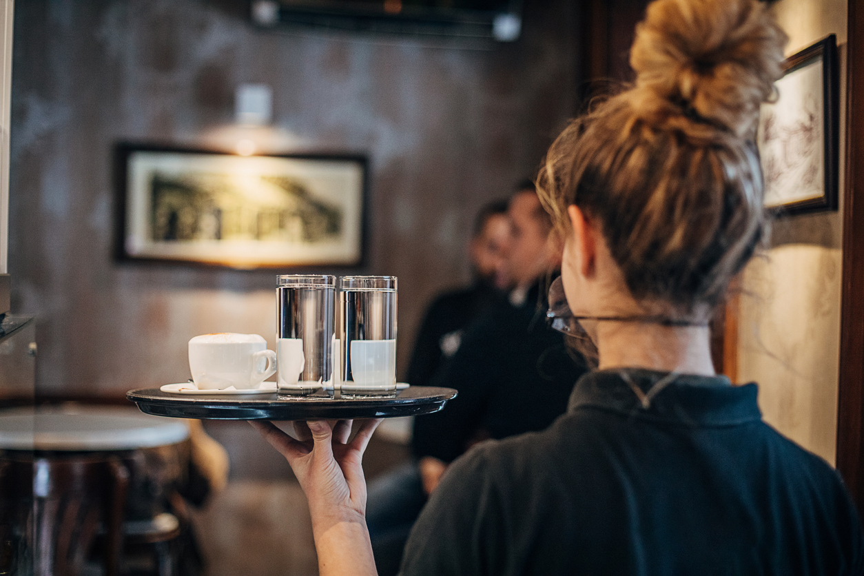 Group of people, female waitress working at cafe, wearing a protective mask. She is holding a trey with coffee cups