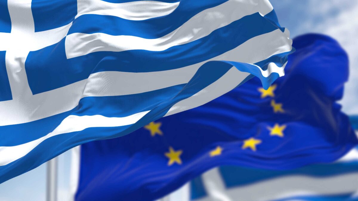 national flag of Greece waving in the wind with blurred european union flag in the background on a clear day