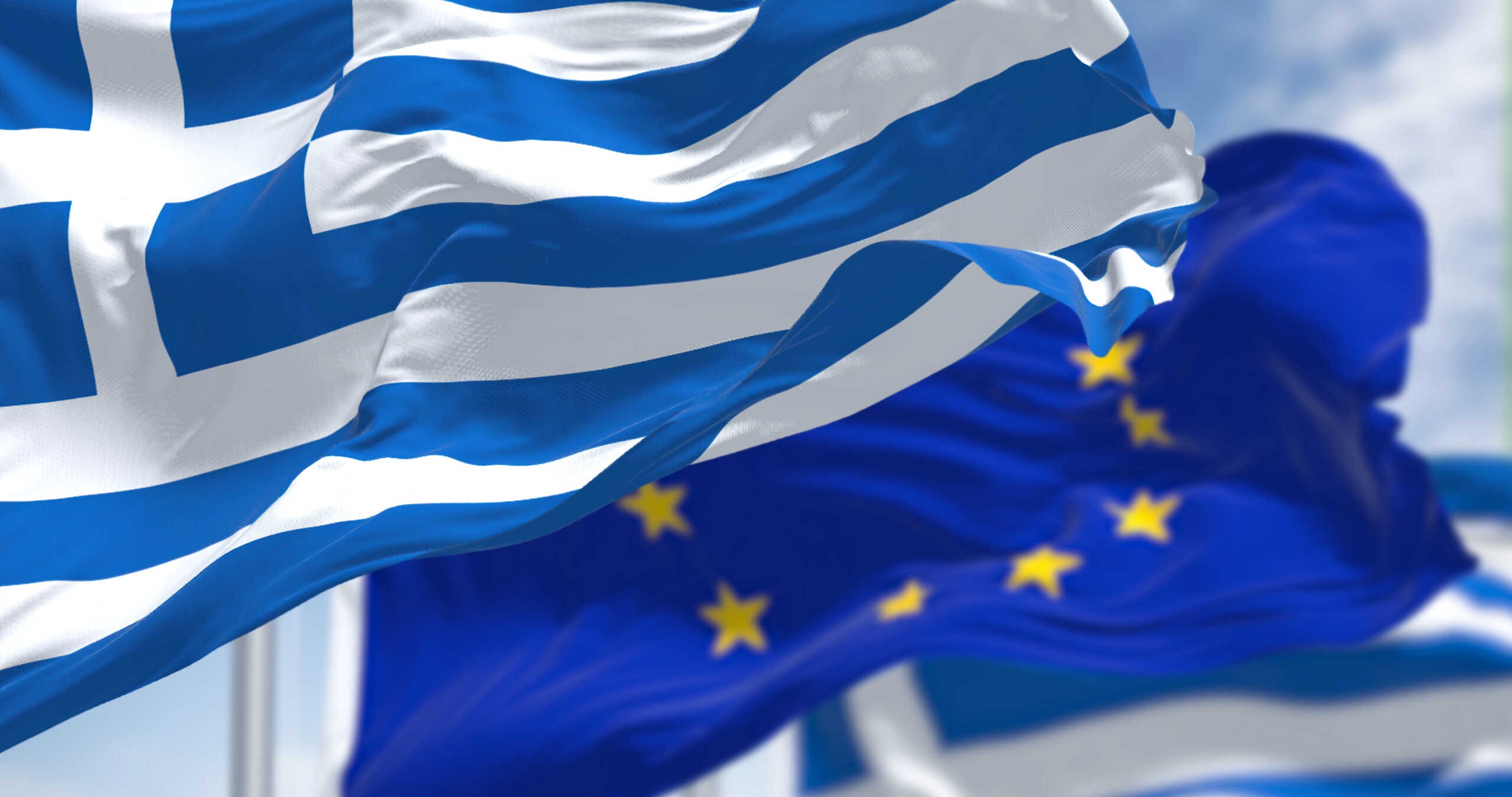 national flag of Greece waving in the wind with blurred european union flag in the background on a clear day