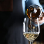Bartender pouring white wine from bottle into glass indoors, closeup. Space for text