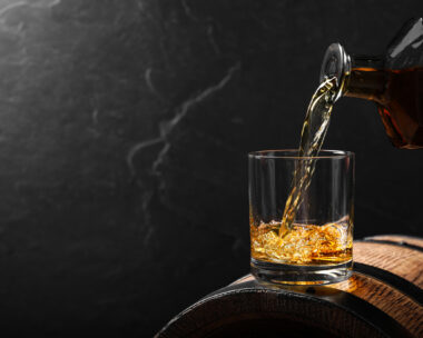 Pouring whiskey into glass from bottle on wooden barrel against grey background, space for text