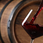 Pouring red wine into glass near wooden barrel, closeup. Space for text