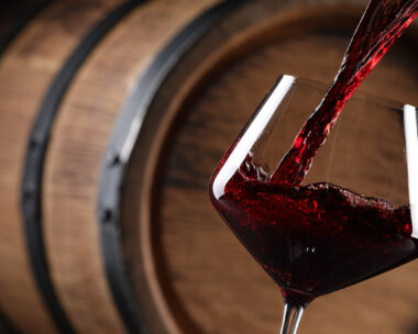 Pouring red wine into glass near wooden barrel, closeup. Space for text