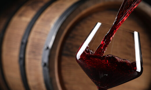 Pouring red wine into glass near wooden barrel, closeup. Space for text