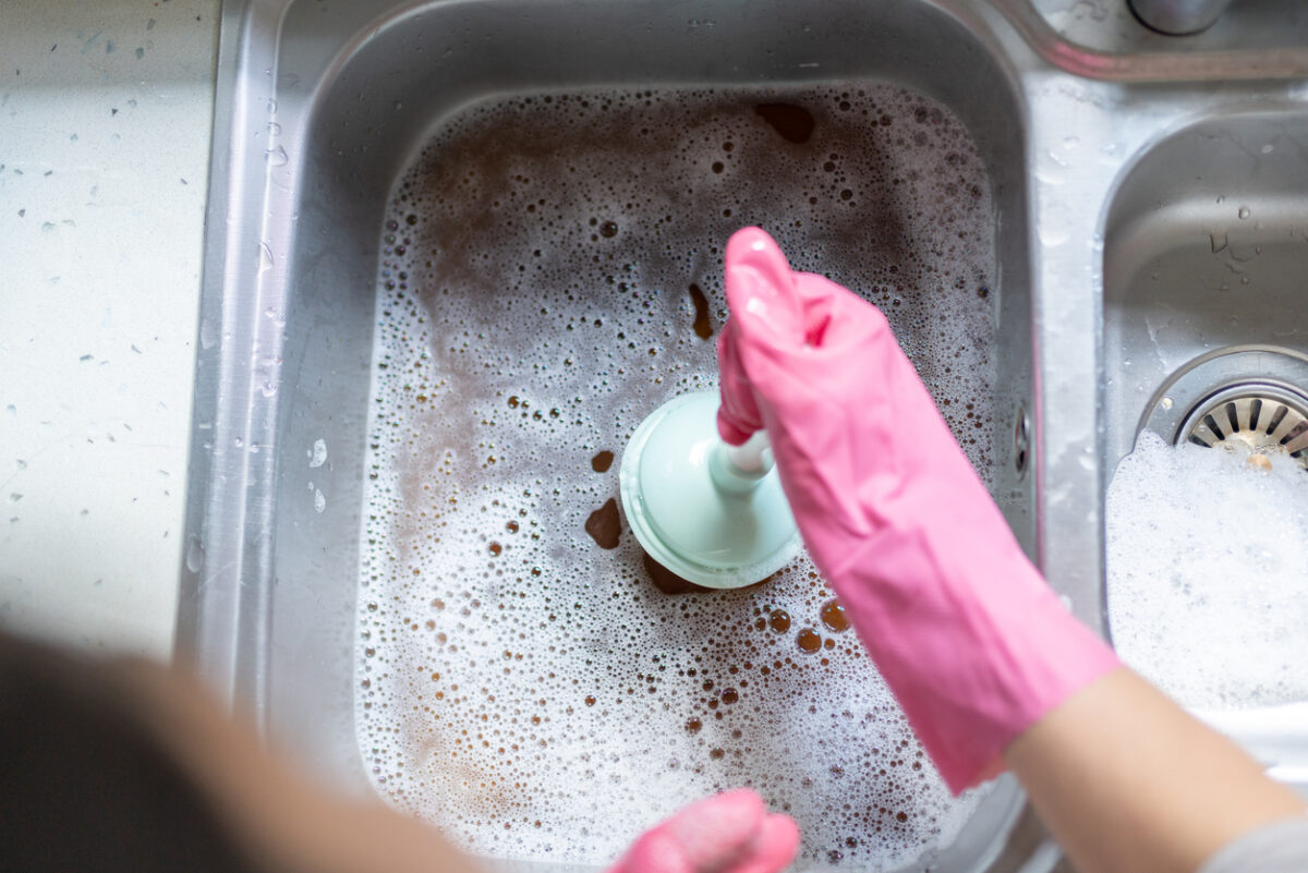 Woman Using Plunger In Blocked Kitchen Sink To Unclog Drain