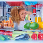 Woman standing in front of opened fridge and holding up to her nose because of bad smell. Picture taken from the inside of fridge full of groceries.