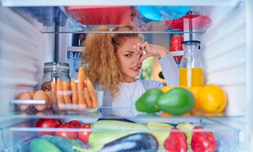 Woman standing in front of opened fridge and holding up to her nose because of bad smell. Picture taken from the inside of fridge full of groceries.