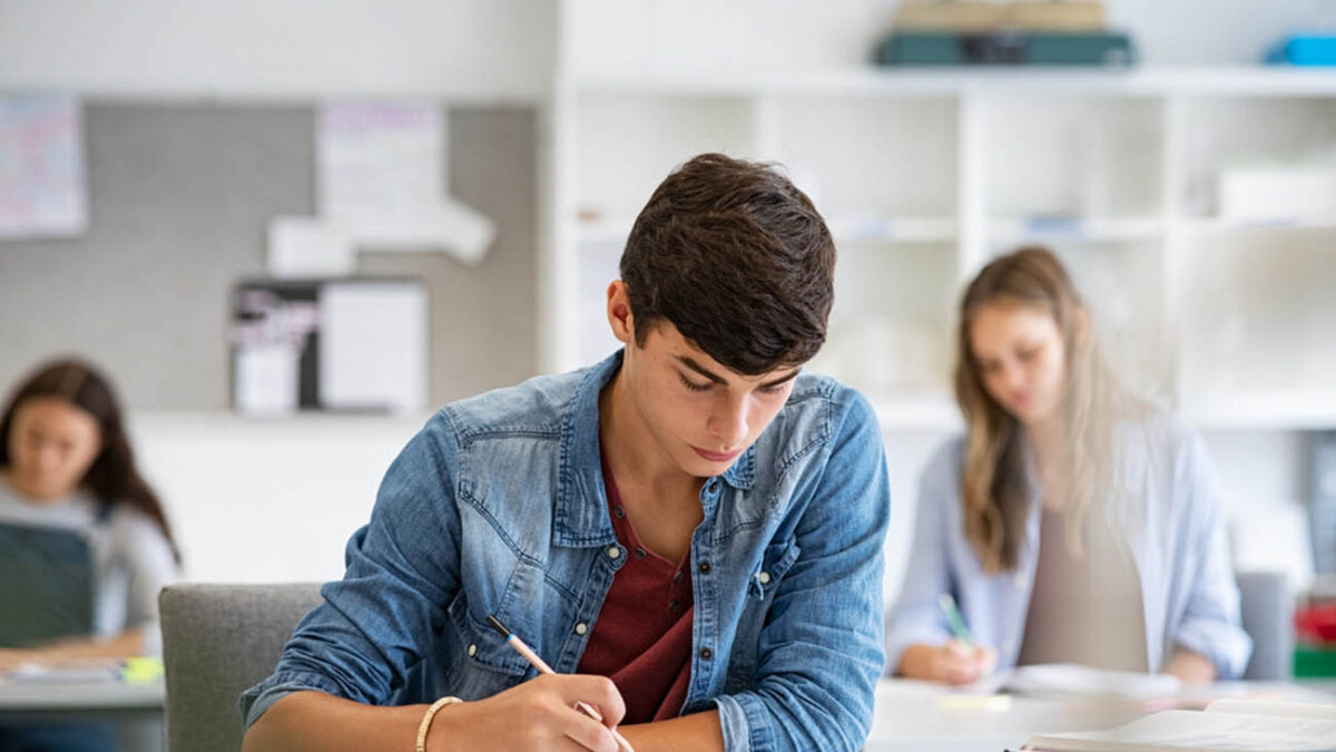Focused young man taking notes from books for his study. College student sitting at desk with books for finding information in high school library. Guy studying in classroom and completing project with highschool classmates in background.