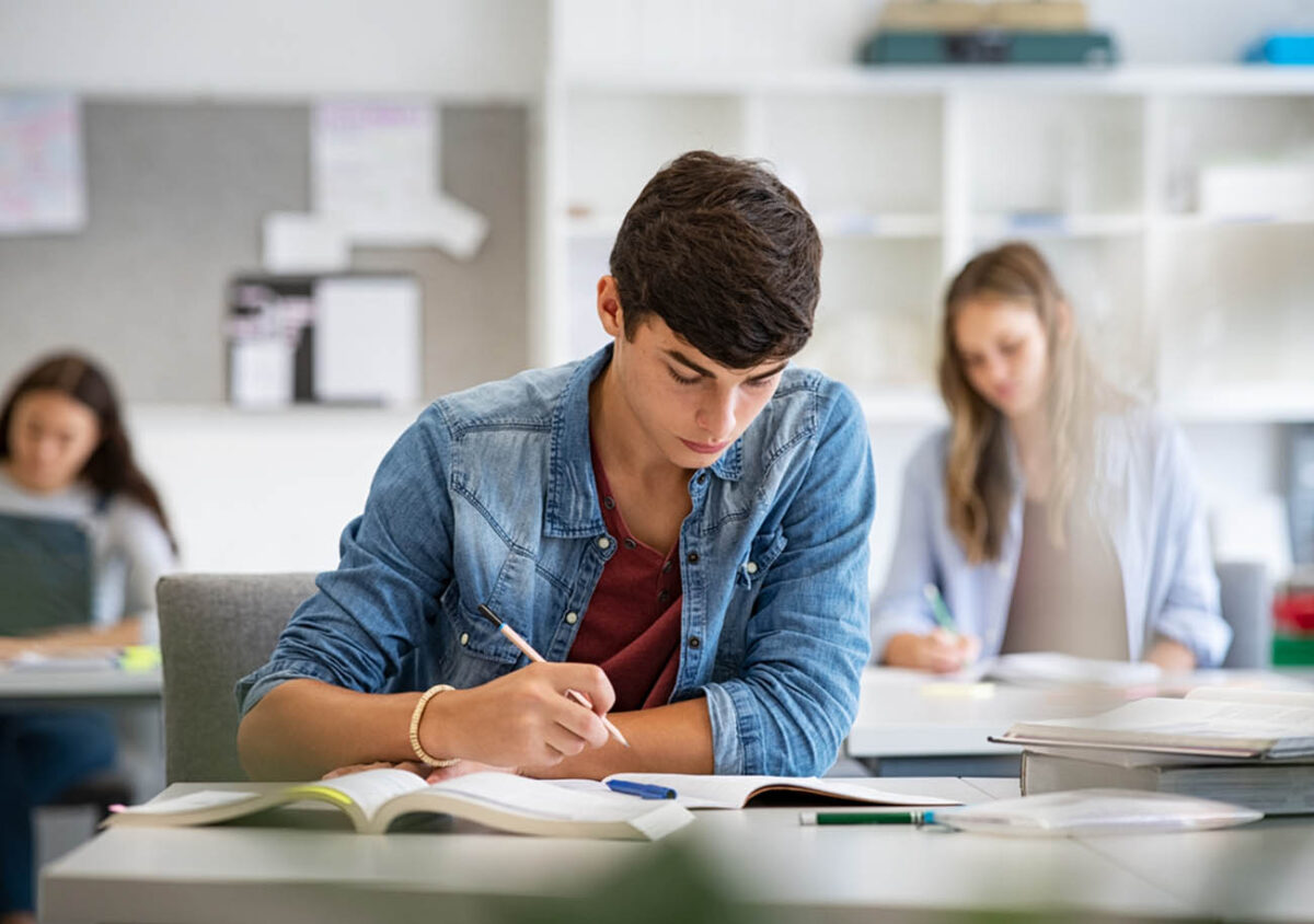 Focused young man taking notes from books for his study. College student sitting at desk with books for finding information in high school library. Guy studying in classroom and completing project with highschool classmates in background.