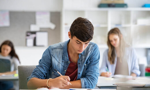 Focused young man taking notes from books for his study. College student sitting at desk with books for finding information in high school library. Guy studying in classroom and completing project with highschool classmates in background.