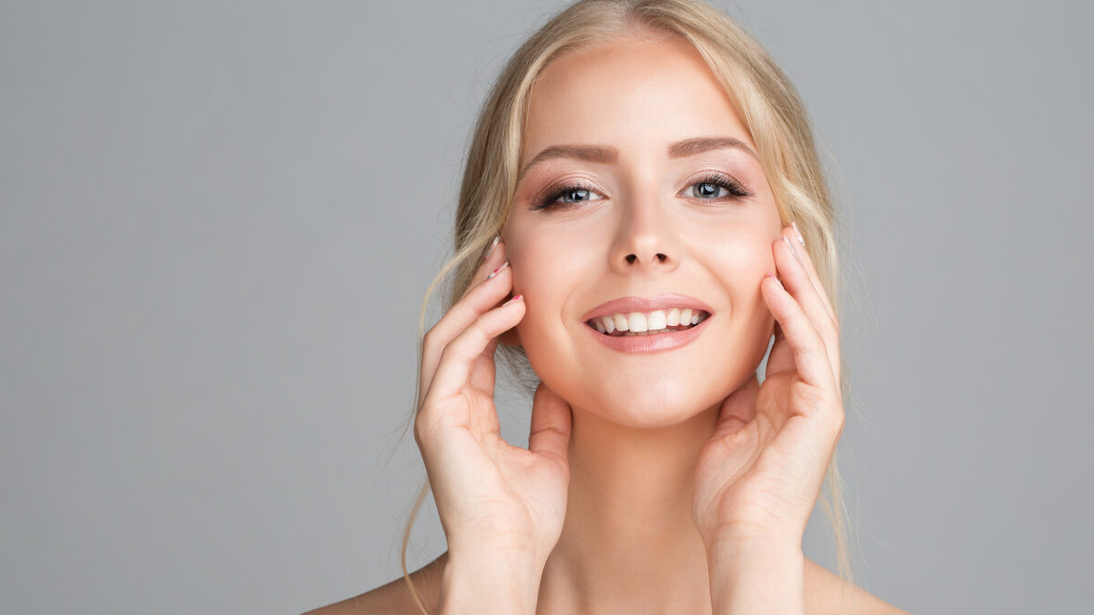 Cheerful Smiling Young Woman Massaging Face Cheek Bones. Beauty Model with Perfect Smooth Skin looking at Camera over Gray Studio Background