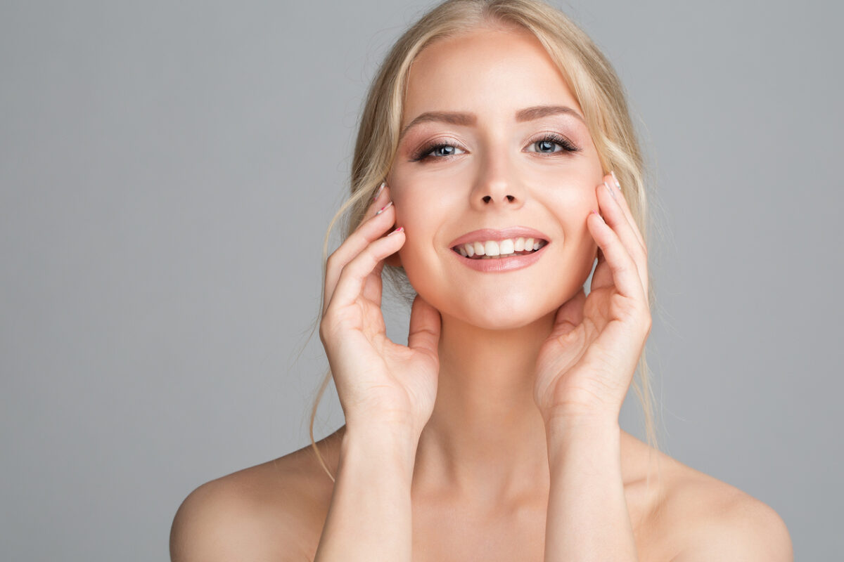 Cheerful Smiling Young Woman Massaging Face Cheek Bones. Beauty Model with Perfect Smooth Skin looking at Camera over Gray Studio Background