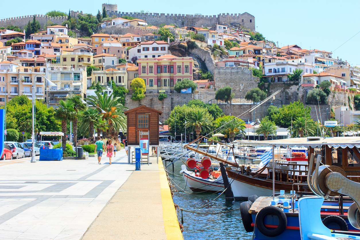 Kavala old town with its Byzantine castle in background, palmtrees, pedestrians on promenade and harbour with fishing boats in foreground. Kavala town,  Makedonia, Greece.